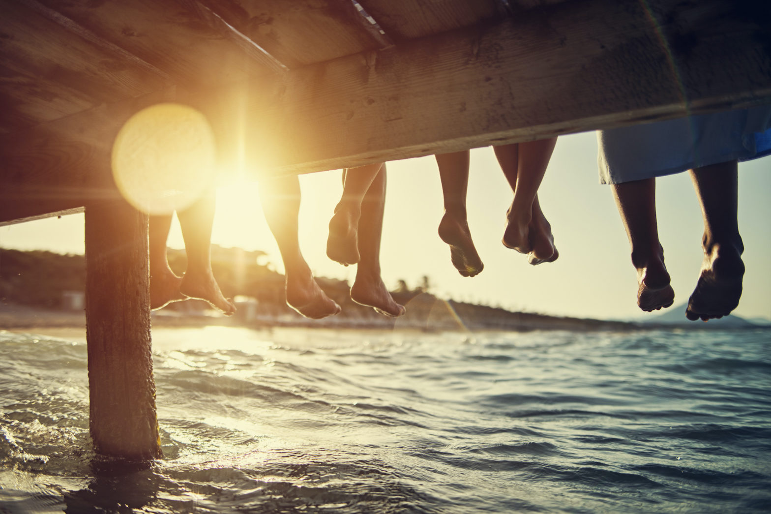 Vijf mensen zitten op de pier. De foto toont hun voeten onder de pier. De zon schijnt onder de pier en onder de voeten door.